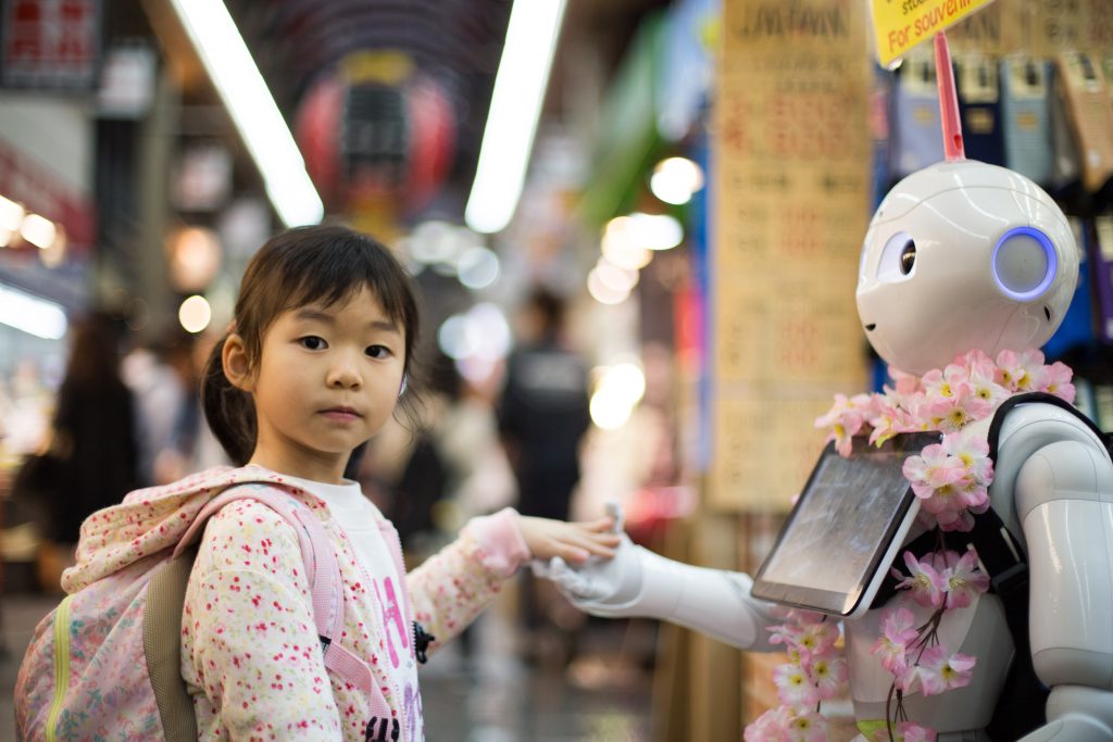 Ein weißer androider Roboter steht in einer Einkaufspassage. Er trägt eine rosa Blumenkette um den Hals und einen Bildschirm auf der Brust. Ihm gegenüber steht ein Kind und reicht ihm die Hand.