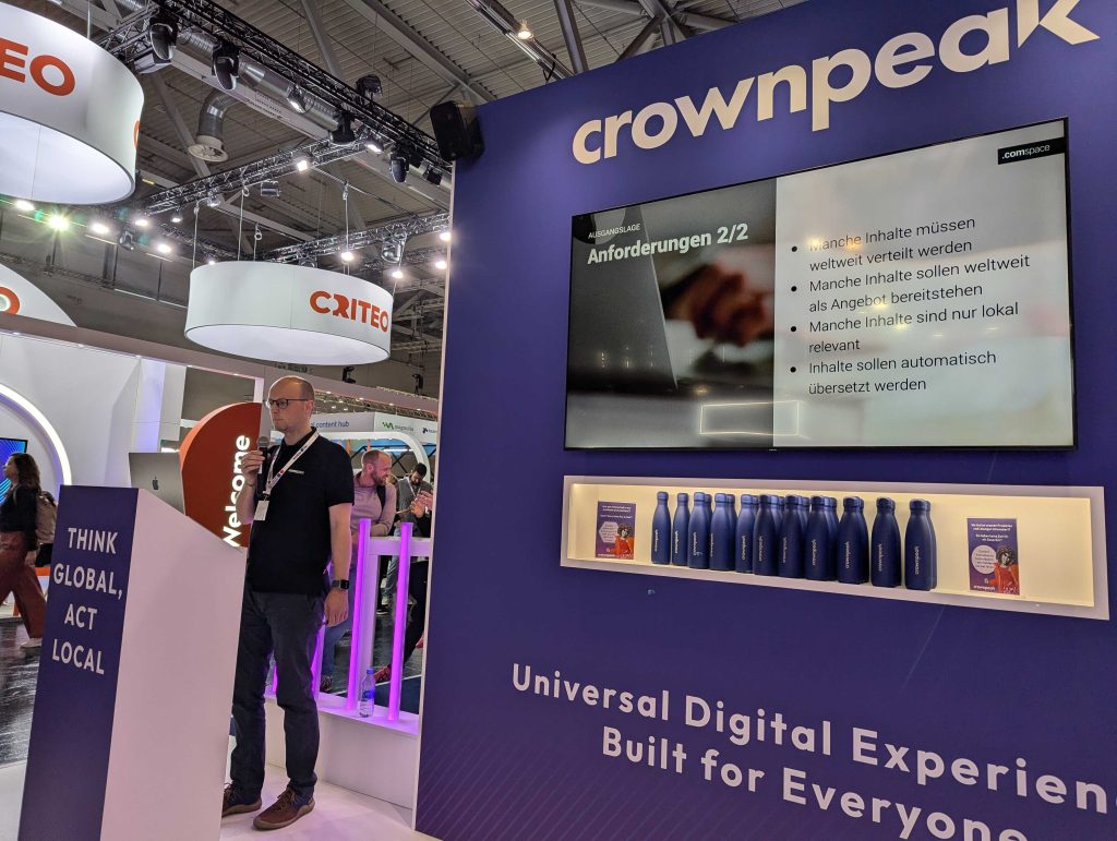 Ein Redner auf der DMEXCO 2024 präsentiert am Crownpeak-Partnerstand vor einer Leinwand mit Anforderungen zur globalen Verteilung und Übersetzung von Inhalten